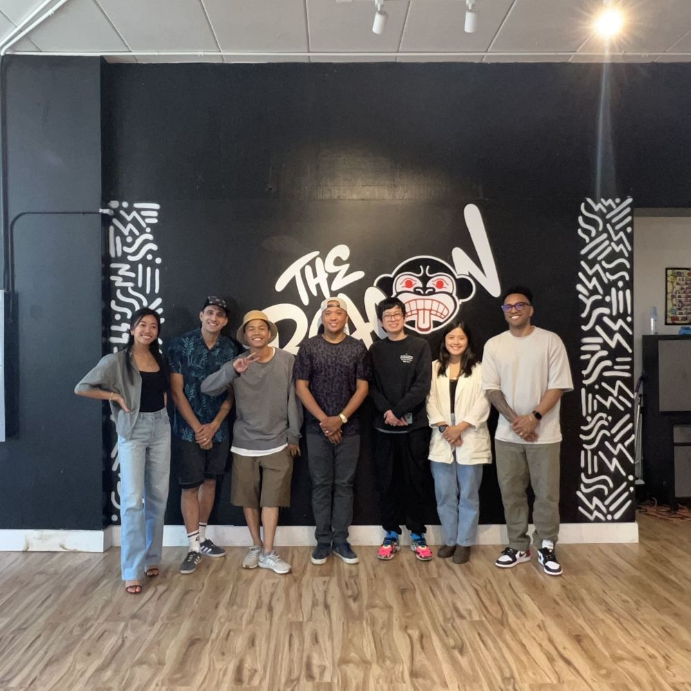 Seven people standing in a row on a light wood floor in front of a dark wall painted with a large graffiti-style mural reading "The Beacon," flanked by black-and-white geometric patterning.