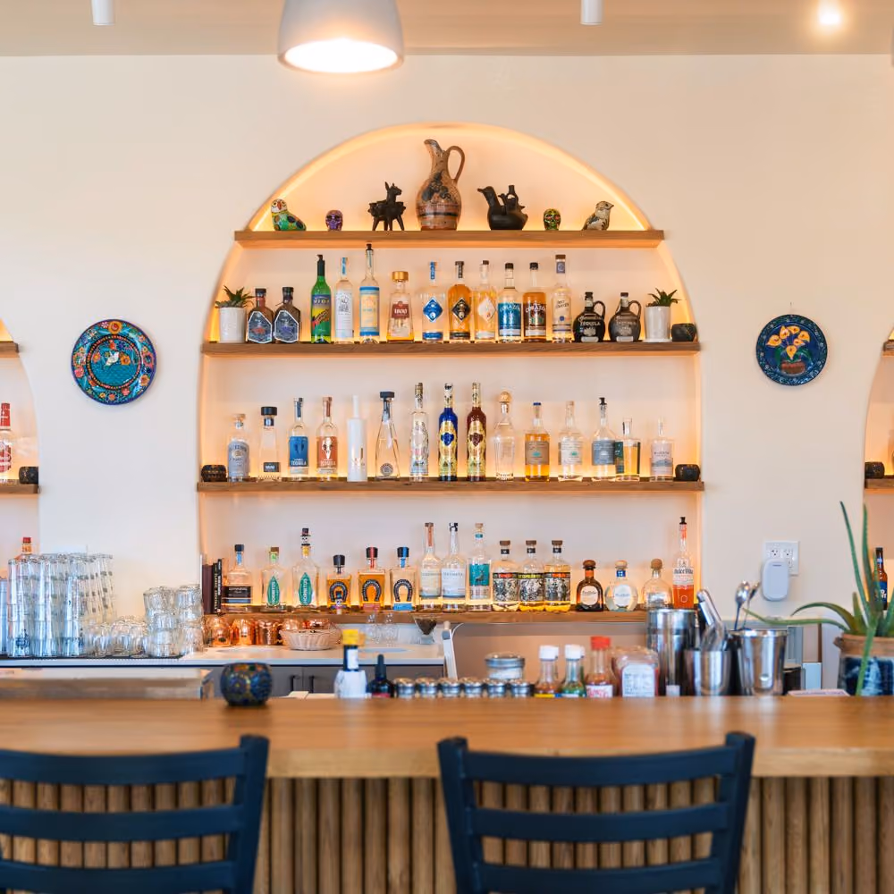 The bar at Baja Bistro: an arched alcove of shelves lined with tequila and mezcal bottles and ceramic figurines, two round painted talavera plates mounted on the white walls, and a wooden bar top with dark blue slatted stools.