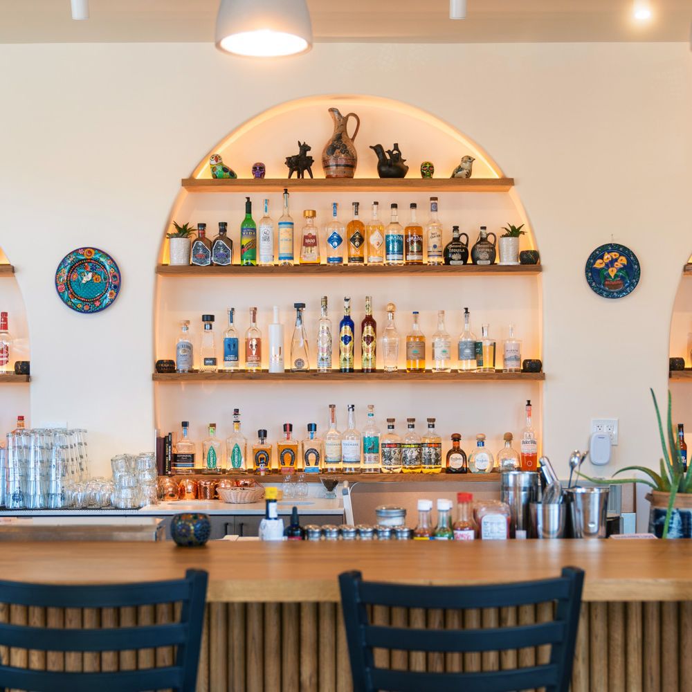 The bar at Baja Bistro: an arched alcove of shelves lined with tequila and mezcal bottles and ceramic figurines, two round painted talavera plates mounted on the white walls, and a wooden bar top with dark blue slatted stools.