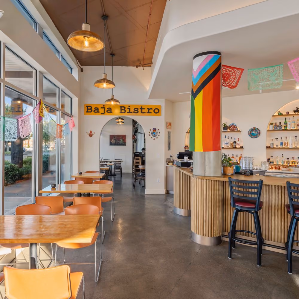The dining room at Baja Bistro, with orange-topped tables and chairs, papel picado banners strung overhead, a structural column wrapped in a Progress Pride flag, a wood-slat service counter, and a "Baja Bistro" sign above an archway at the back.