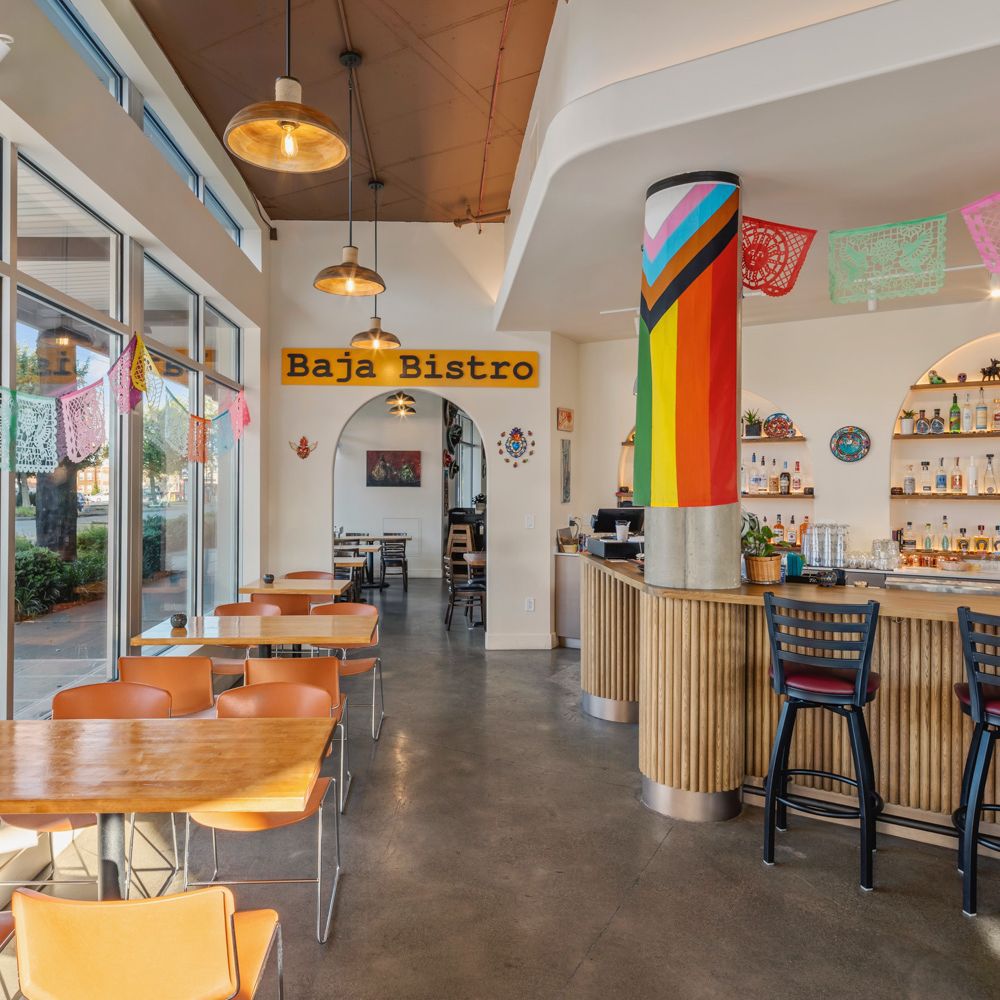 The dining room at Baja Bistro, with orange-topped tables and chairs, papel picado banners strung overhead, a structural column wrapped in a Progress Pride flag, a wood-slat service counter, and a "Baja Bistro" sign above an archway at the back.