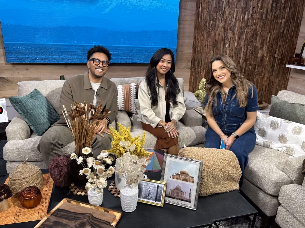 Three people seated on a sectional sofa on the set of A New Day Northwest, with a coffee table in front of them styled with dried florals in vases, framed photographs, a wooden serving board, and textured decorative pumpkins; a large screen showing water and a wood-slat wall form the backdrop.