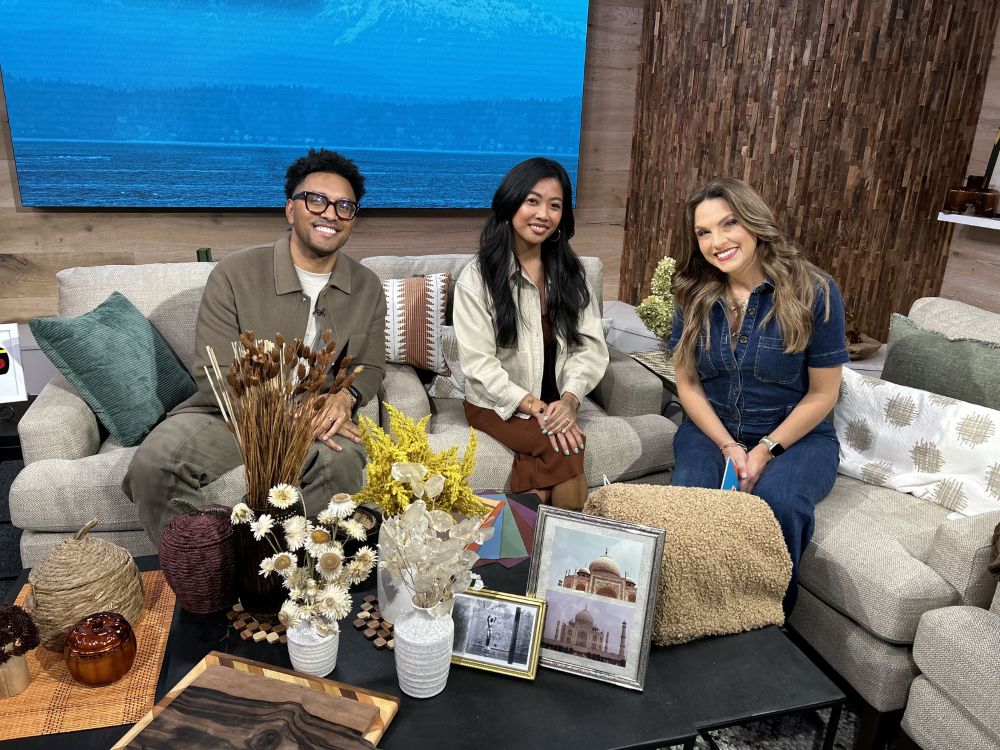 Three people seated on a sectional sofa on the set of A New Day Northwest, with a coffee table in front of them styled with dried florals in vases, framed photographs, a wooden serving board, and textured decorative pumpkins; a large screen showing water and a wood-slat wall form the backdrop.