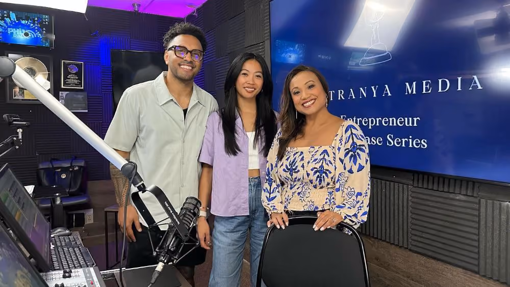 Three people standing together in a recording studio, with a boom microphone arm in the foreground, a mixing console and framed plaques to the side, and a blue screen behind them reading "Patranya Media — Entrepreneur Showcase Series."