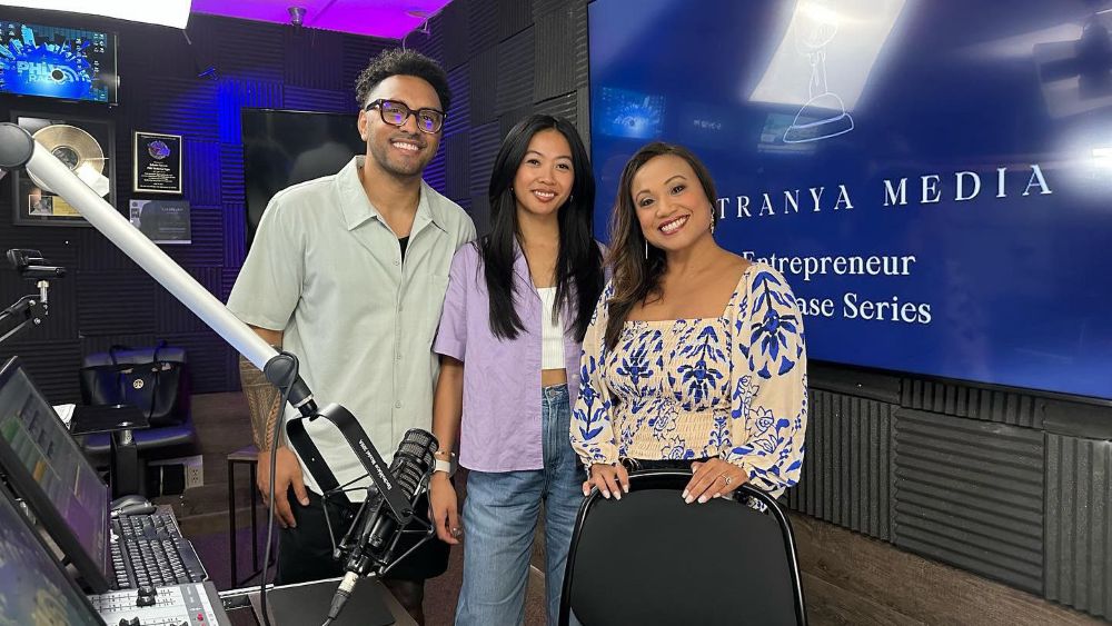 Three people standing together in a recording studio, with a boom microphone arm in the foreground, a mixing console and framed plaques to the side, and a blue screen behind them reading "Patranya Media — Entrepreneur Showcase Series."