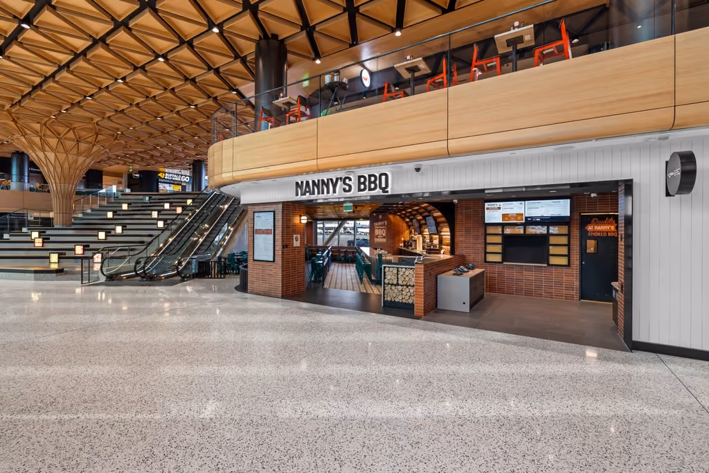 The Nanny's BBQ storefront seen from across the concourse at SEA airport, with its name on a sign above the entrance, an orange brick facade, digital menu boards, and a sweeping timber ceiling and escalators to the left.