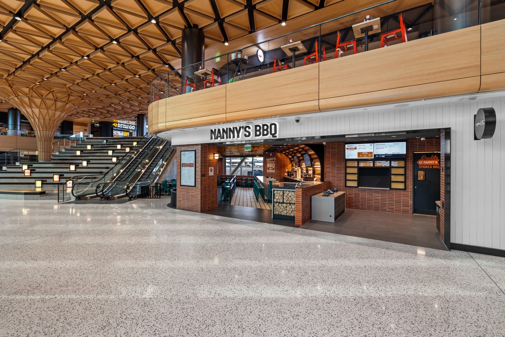The Nanny's BBQ storefront seen from across the concourse at SEA airport, with its name on a sign above the entrance, an orange brick facade, digital menu boards, and a sweeping timber ceiling and escalators to the left.