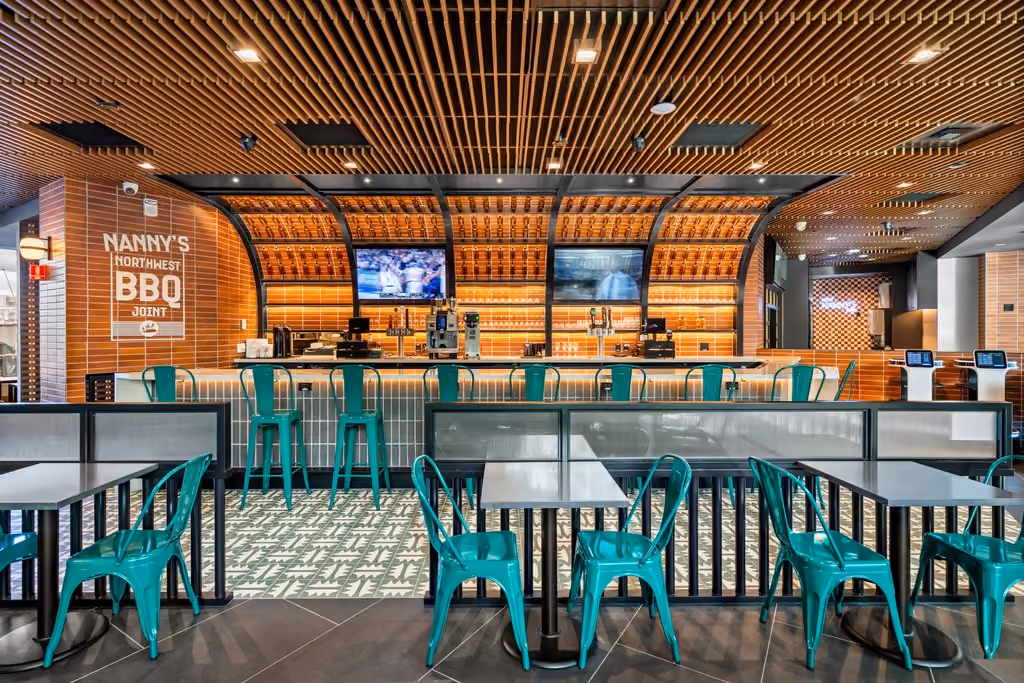 The bar at Nanny's BBQ, with arched orange-tiled vaults over backlit bottle shelving, two televisions, teal metal bar stools along a tiled counter, and cafe tables with teal chairs in the foreground.