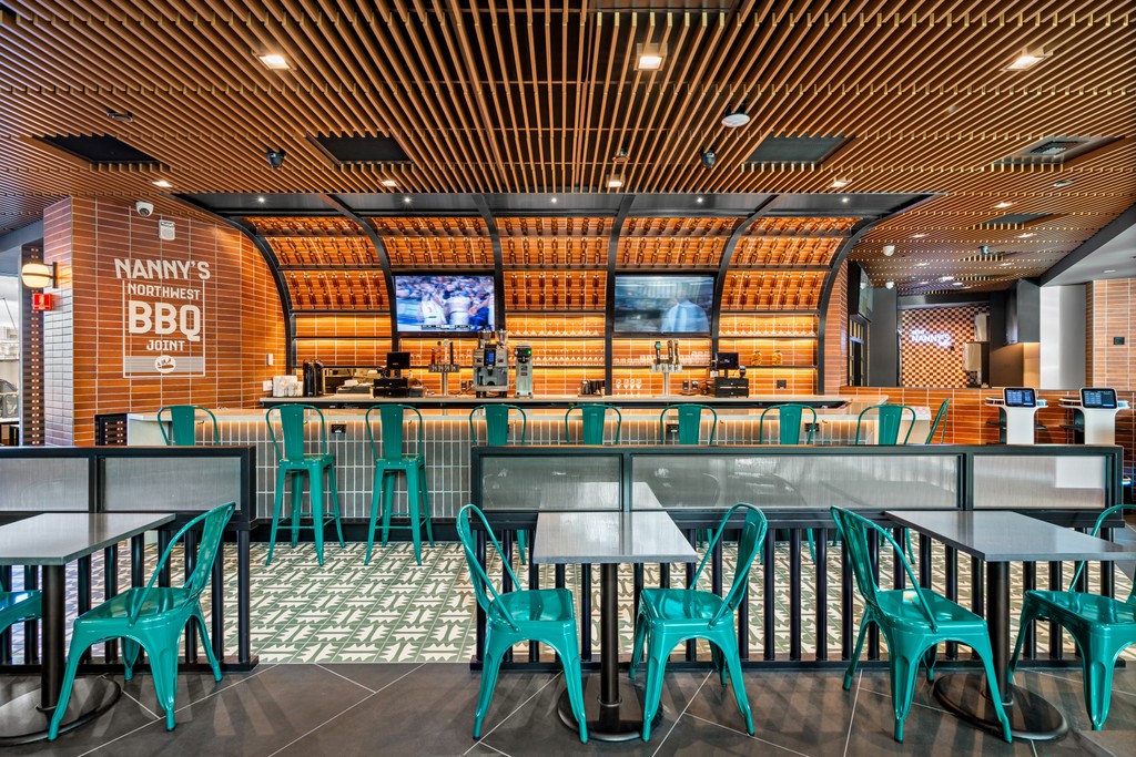 The bar at Nanny's BBQ, with arched orange-tiled vaults over backlit bottle shelving, two televisions, teal metal bar stools along a tiled counter, and cafe tables with teal chairs in the foreground.