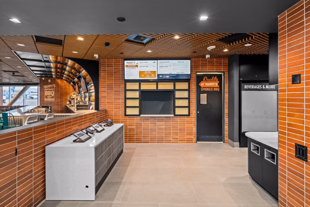 The order area at Nanny's BBQ: a stainless steel service counter, a wall of orange brick tile, digital menu screens, a beverage station, and a slatted wood ceiling overhead.
