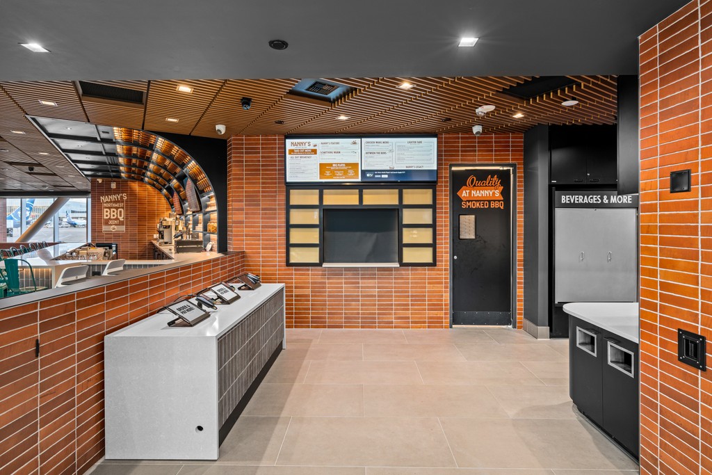 The order area at Nanny's BBQ: a stainless steel service counter, a wall of orange brick tile, digital menu screens, a beverage station, and a slatted wood ceiling overhead.