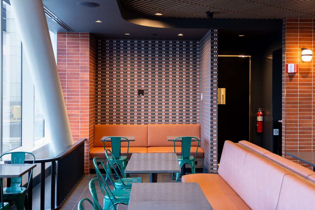 A quieter seating nook at Nanny's BBQ with pink upholstered banquettes, small tables and teal chairs, a patterned dark tile feature wall, and a curved window looking out to the airfield.