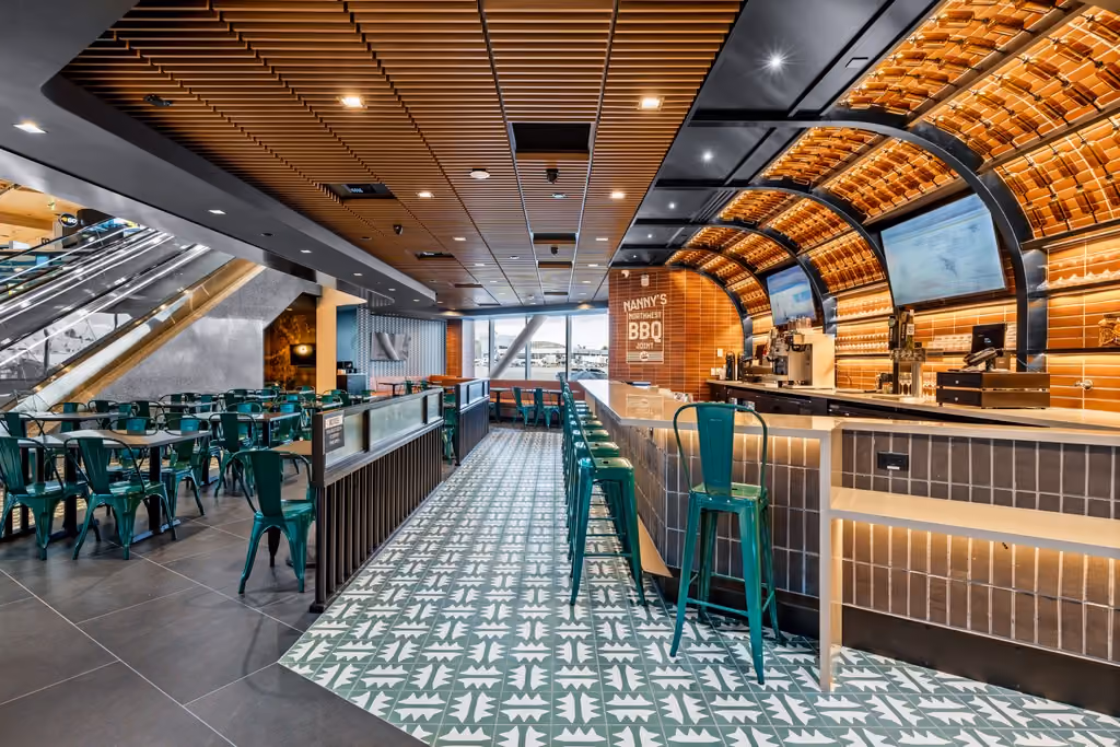 A wide view down the length of Nanny's BBQ, showing the patterned tile floor, rows of teal chairs and tables, the bar counter with under-lit tile, and the arched timber-lined ceiling above.