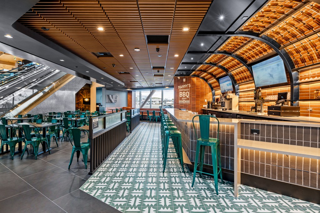 A wide view down the length of Nanny's BBQ, showing the patterned tile floor, rows of teal chairs and tables, the bar counter with under-lit tile, and the arched timber-lined ceiling above.