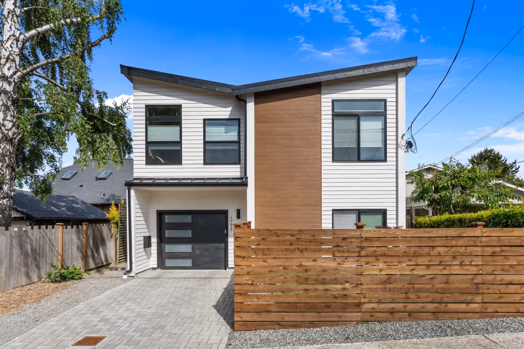 The completed North Admiral detached accessory dwelling unit: a two-story home with white horizontal siding and a warm wood-clad center bay, a dark garage door, paver driveway, and a horizontal cedar fence, under a clear blue sky.