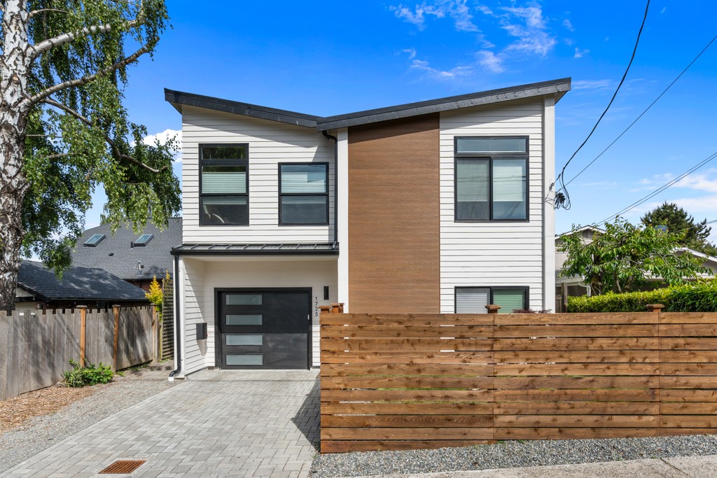 The completed North Admiral detached accessory dwelling unit: a two-story home with white horizontal siding and a warm wood-clad center bay, a dark garage door, paver driveway, and a horizontal cedar fence, under a clear blue sky.