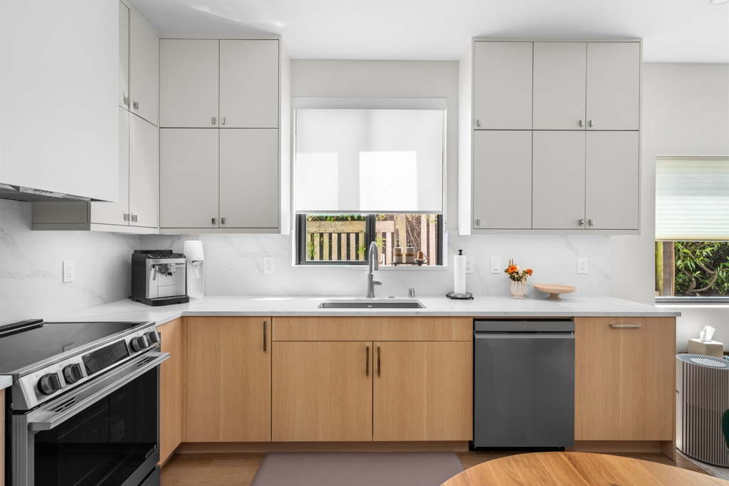 The DADU kitchen with pale wood lower cabinets, light grey uppers, white stone counters and backsplash, a stainless range and dishwasher, and a window above the sink looking onto greenery.