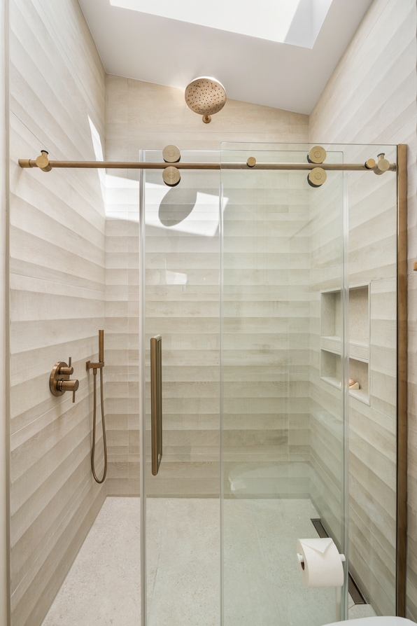 A walk-in shower with large-format cream tile, a sliding glass door and brushed brass fixtures and hardware, a recessed niche, and daylight from a skylight above.