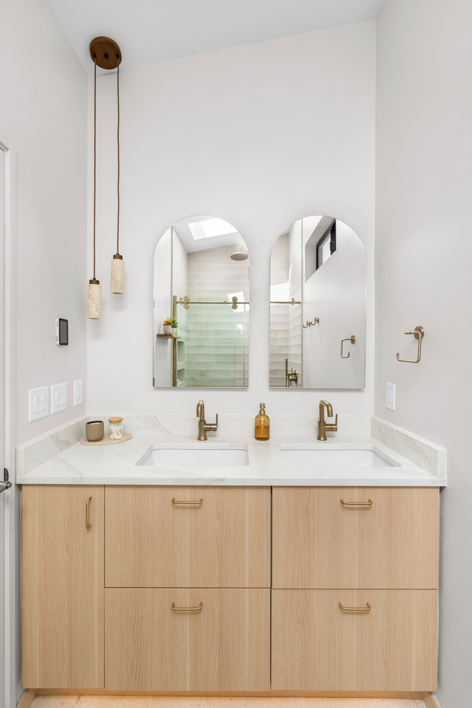 A double vanity with pale wood drawers and brass pulls, a white stone counter, two arched mirrors, brass faucets, and slender pendant lights hanging on either side.