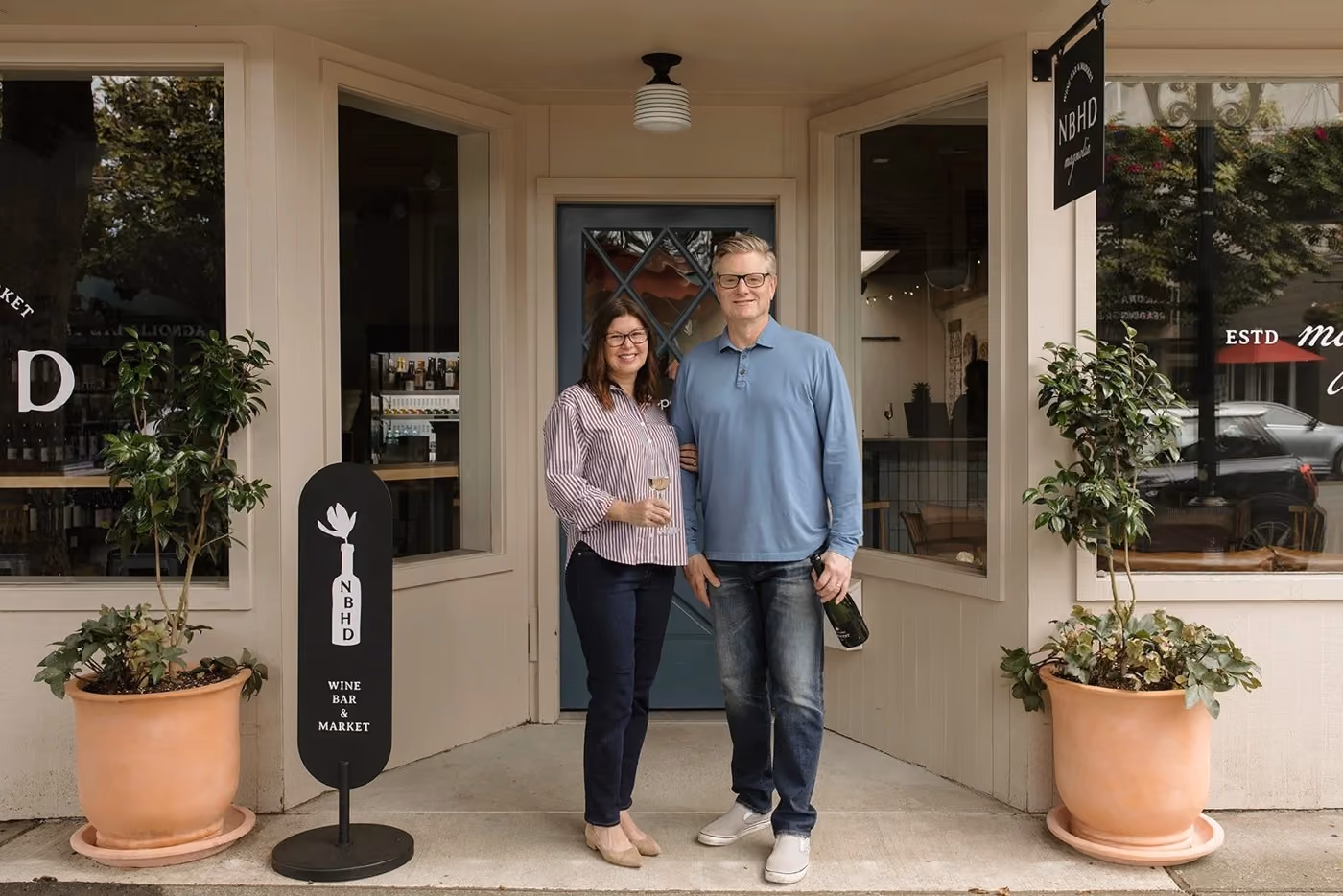 Teddi and Patrick standing together in the doorway of NBHD Wine & Market, one holding a glass and the other a bottle, flanked by potted trees in terracotta planters and a black sidewalk sign with the shop's logo.