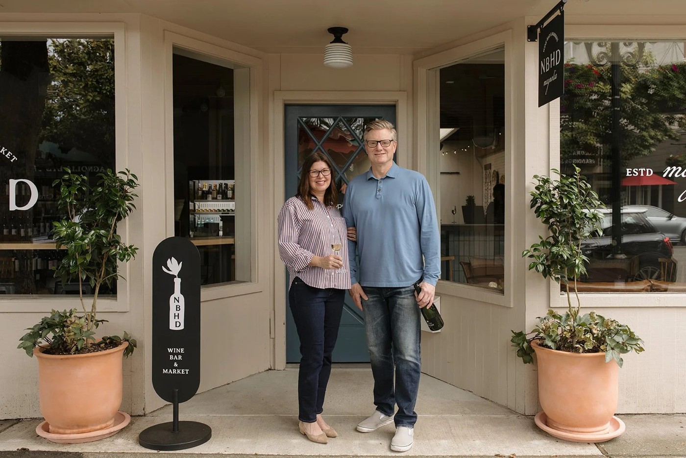 Teddi and Patrick standing together in the doorway of NBHD Wine & Market, one holding a glass and the other a bottle, flanked by potted trees in terracotta planters and a black sidewalk sign with the shop's logo.
