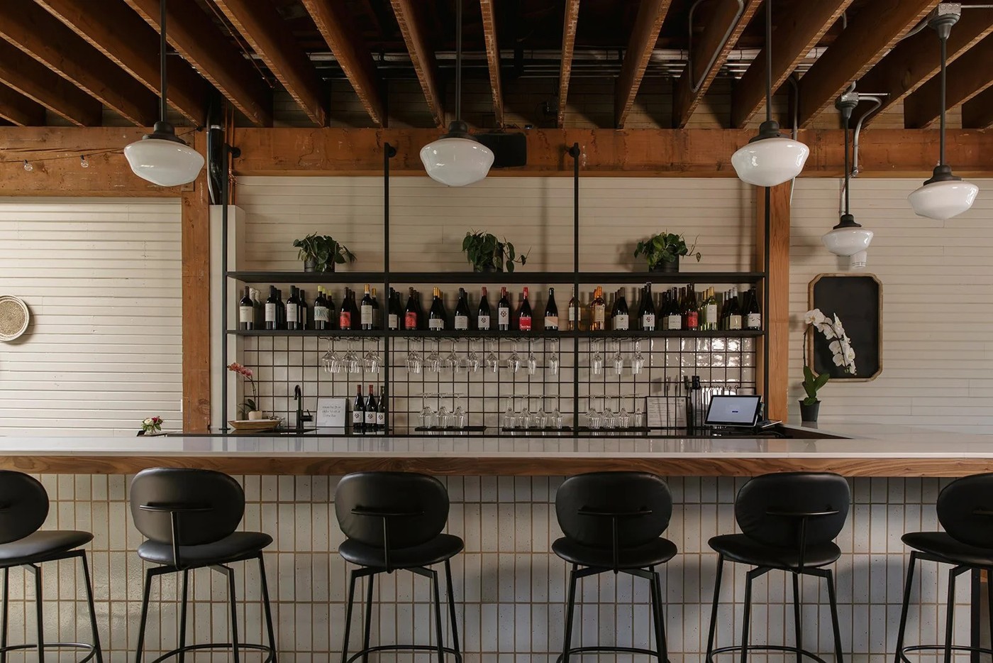The bar at NBHD Wine & Market: black leather stools along a tiled counter with a wood top, a black metal shelving unit holding rows of wine bottles and hanging glassware, plants above, and white schoolhouse pendant lights beneath exposed timber beams.