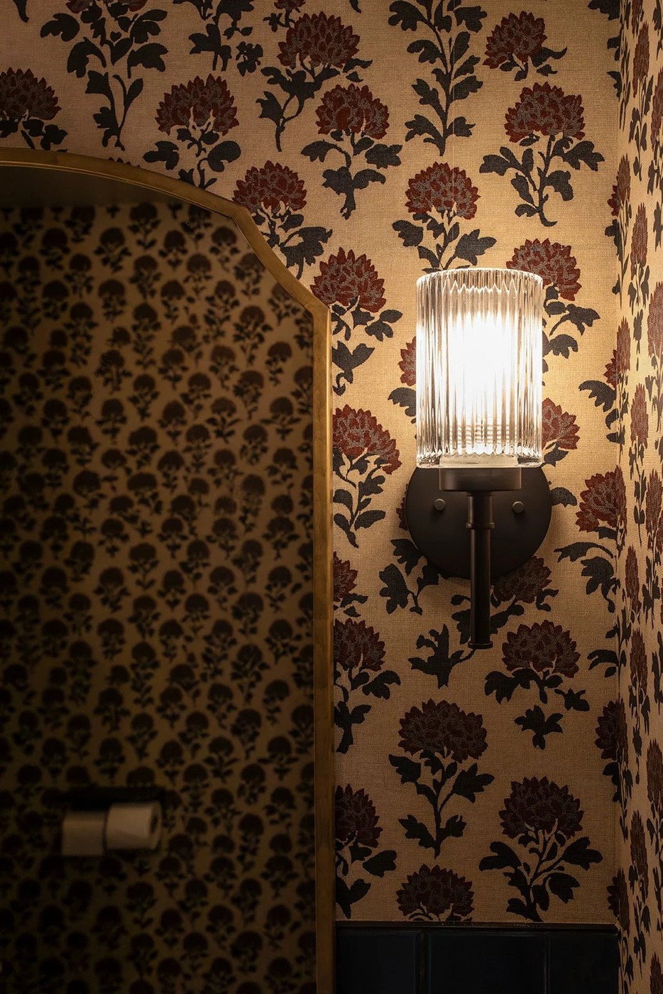 A detail of the restroom at NBHD Wine & Market, with dark floral block-print wallpaper on a warm gold ground and a fluted glass wall sconce casting a soft glow.
