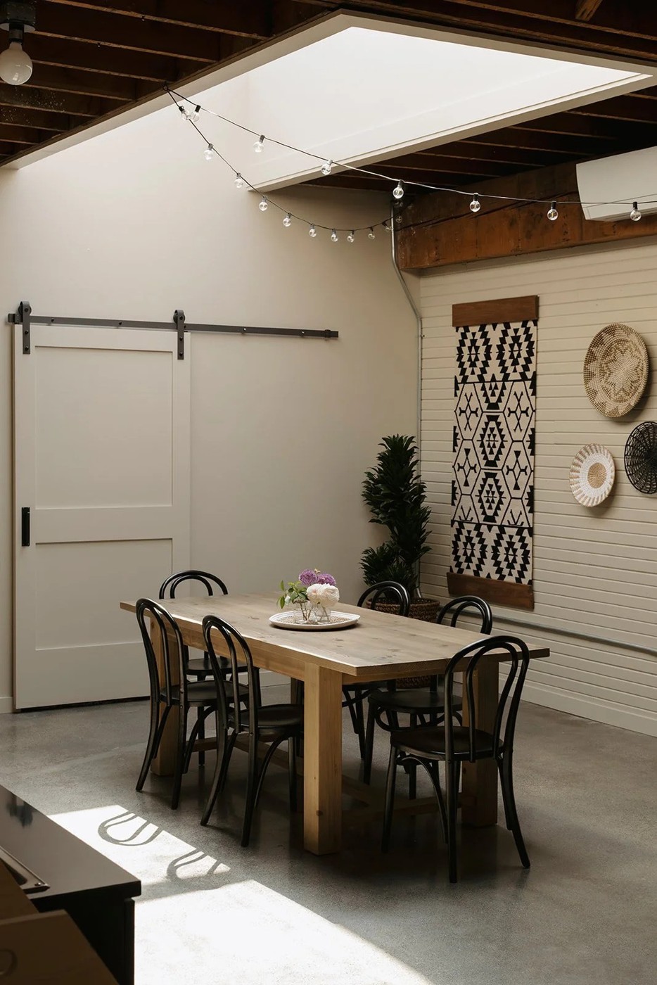 A covered patio with a long wood table and black bentwood chairs, a sliding barn door, string lights overhead, and a wall hung with woven baskets and a patterned textile.