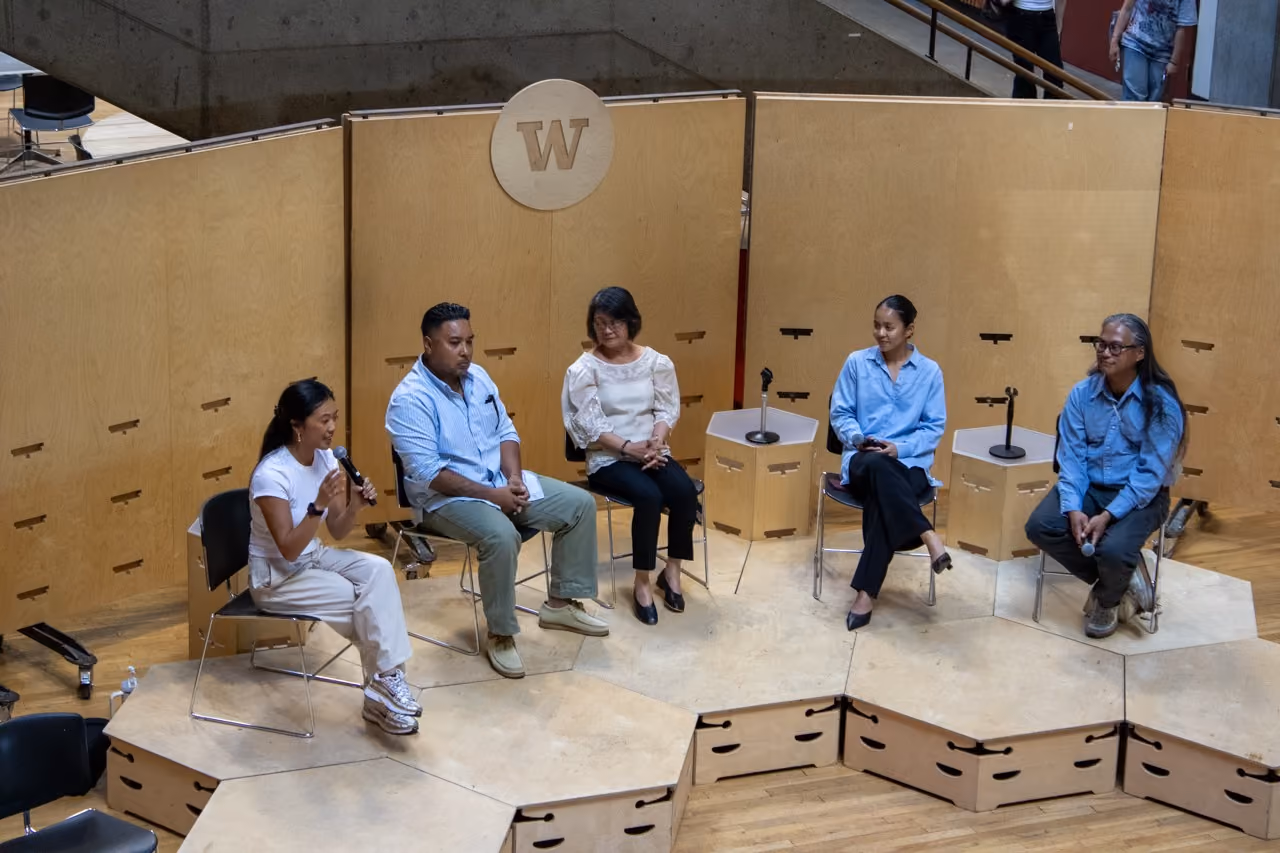 Five panelists seated on a low wooden stage in front of a plywood backdrop bearing the University of Washington W, one speaking into a handheld microphone, during the Fil-Am sa CBE panel.