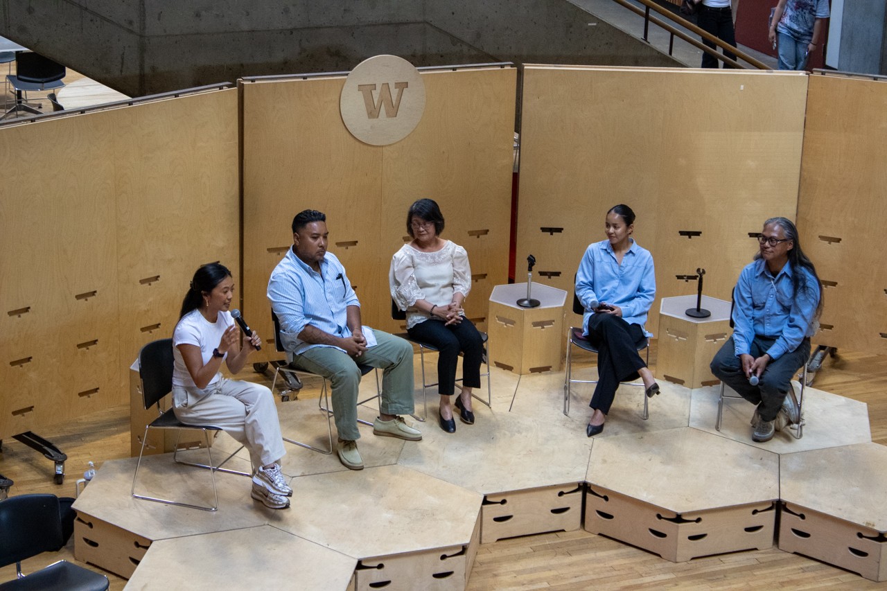 Five panelists seated on a low wooden stage in front of a plywood backdrop bearing the University of Washington W, one speaking into a handheld microphone, during the Fil-Am sa CBE panel.