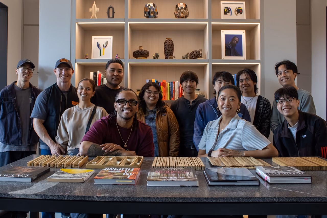 A large group of students and studio members gathered for a group photo behind a stone-topped table spread with design books and material samples, in front of shelves displaying objects, artwork, and books at the Mount Baker studio.