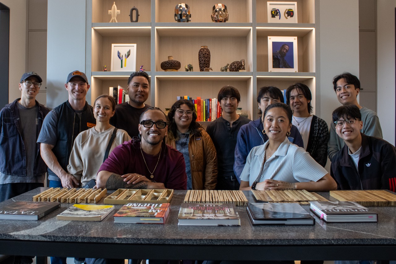 A large group of students and studio members gathered for a group photo behind a stone-topped table spread with design books and material samples, in front of shelves displaying objects, artwork, and books at the Mount Baker studio.