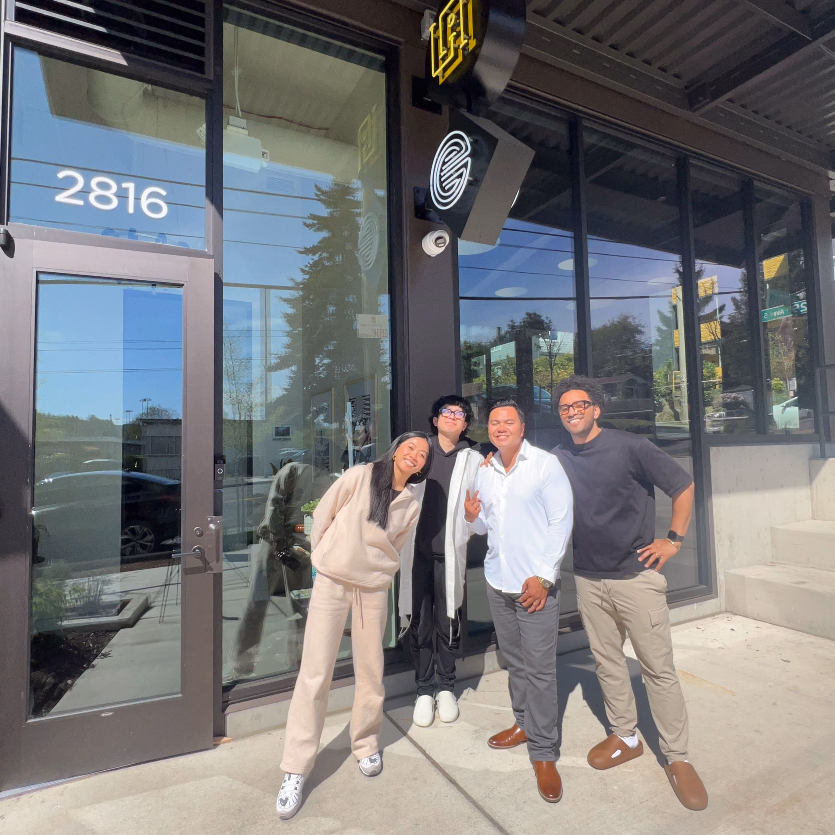 Four members of the La Union Studio team standing together and smiling outside the studio's glass storefront entrance, numbered 2816, on a sunny day.