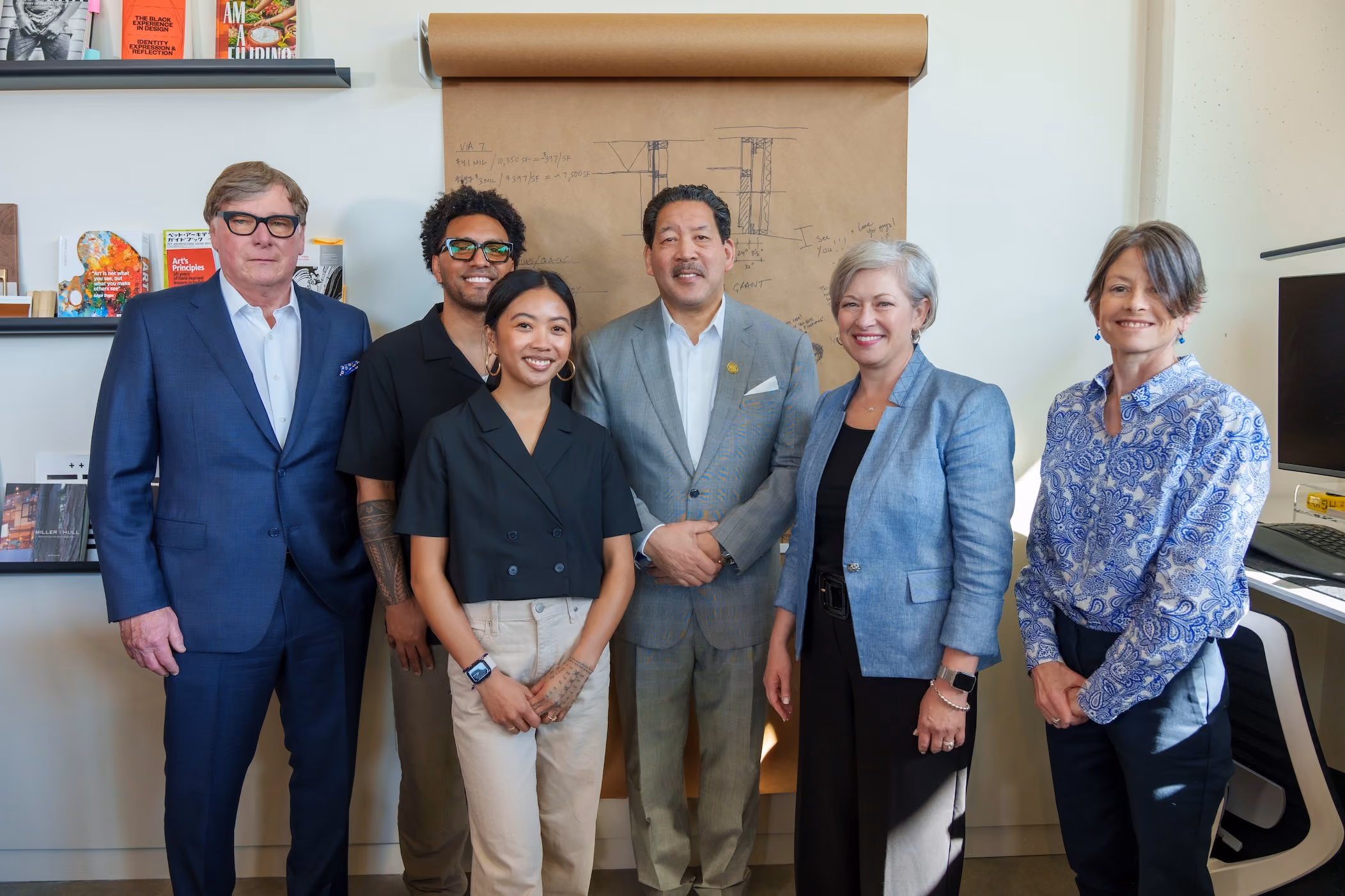 Six people standing shoulder to shoulder inside the La Union Studio office, in front of a wall pinned with a large architectural drawing and shelves of books and objects.
