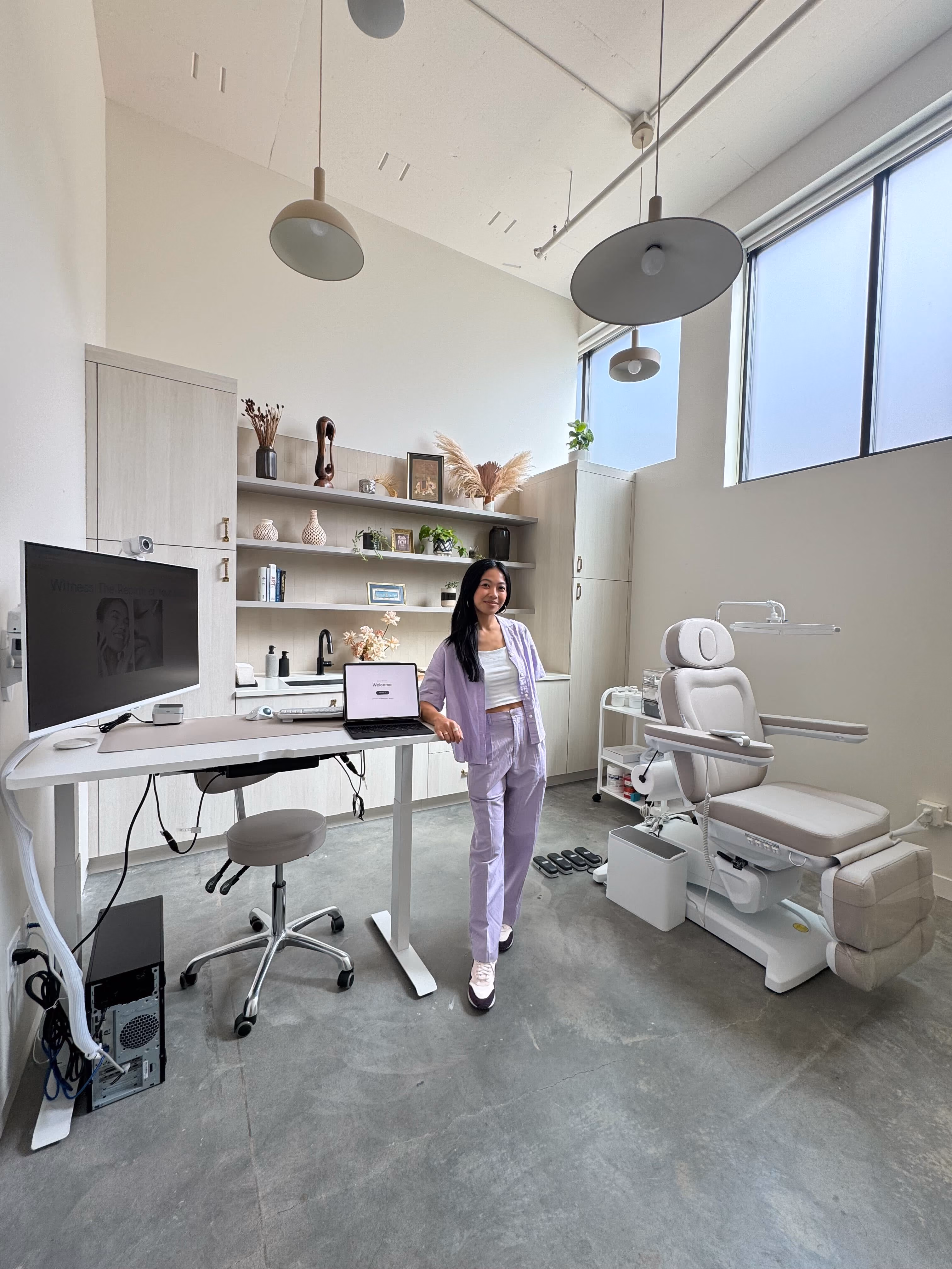 Sonia-Lynn Abenojar standing in the Emblem Skincare treatment room, beside a desk and a reclining treatment chair, with shelves of skincare products and pendant lamps overhead.