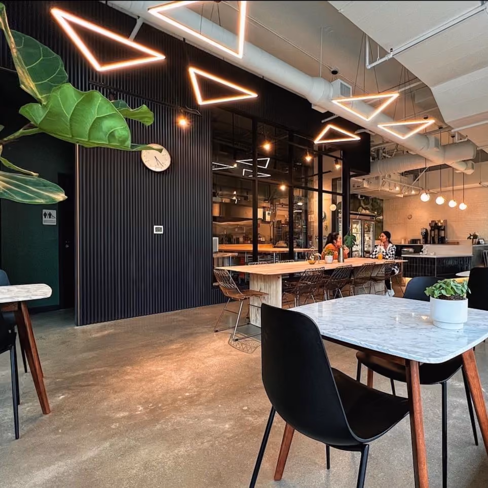 The seating area at Raised Doughnuts & Cakes, with angular neon triangle light fixtures suspended from the ceiling, dark slatted walls, marble-topped tables, black chairs, and a hanging plant.