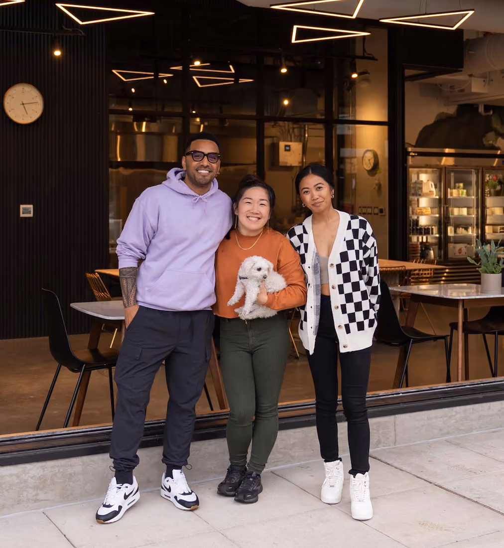 Three people standing in front of the Raised Doughnuts & Cakes storefront window at night, one of them holding a small white dog.