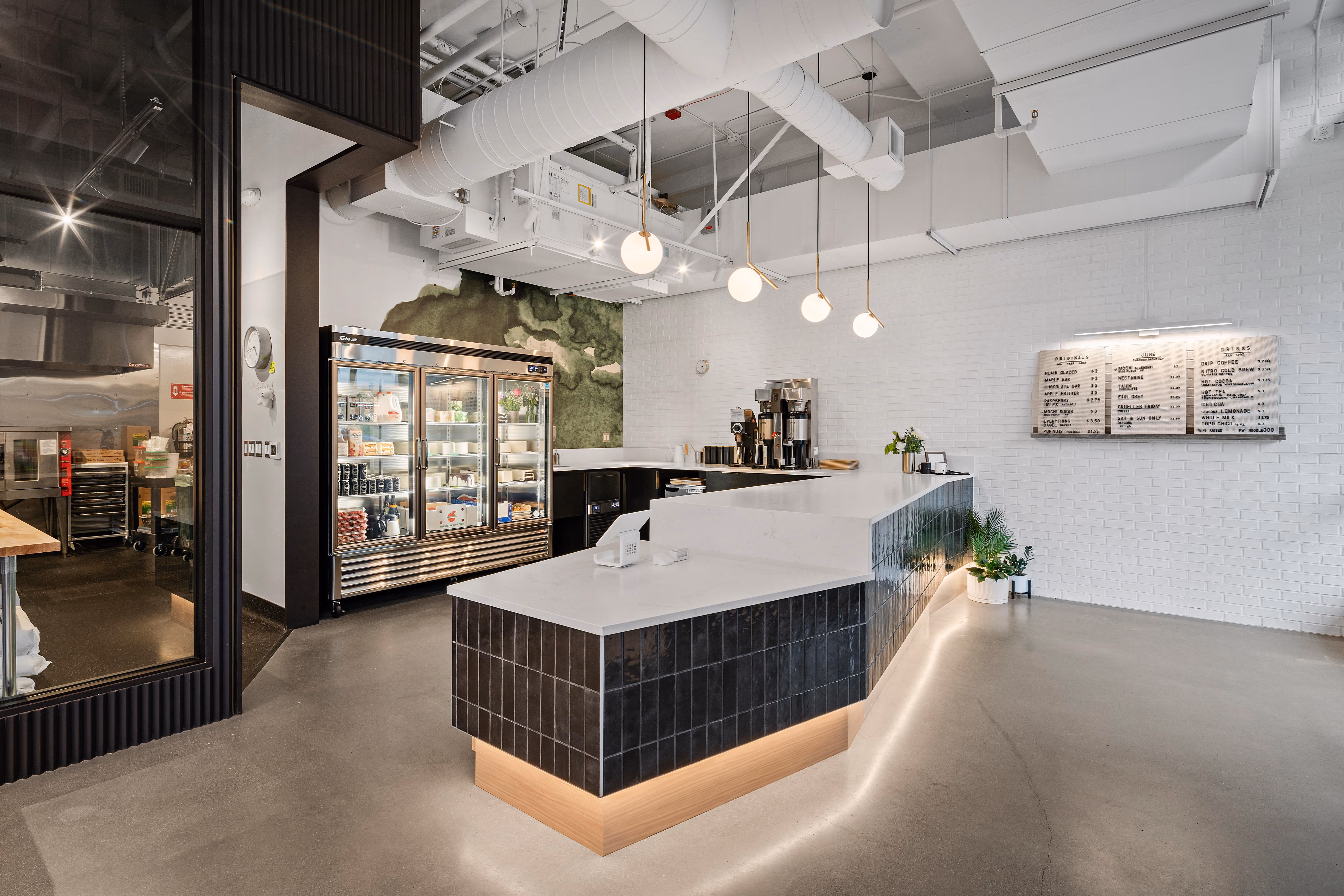 The service counter at Raised Doughnuts & Cakes: a curved dark-tiled counter with a white stone top and illuminated base, a glass pastry case, exposed white ductwork, white brick walls, and menu boards.