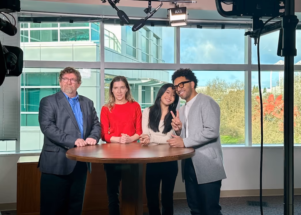Four people standing around a round table in a television studio, with lighting rigs overhead and windows looking onto trees behind them.