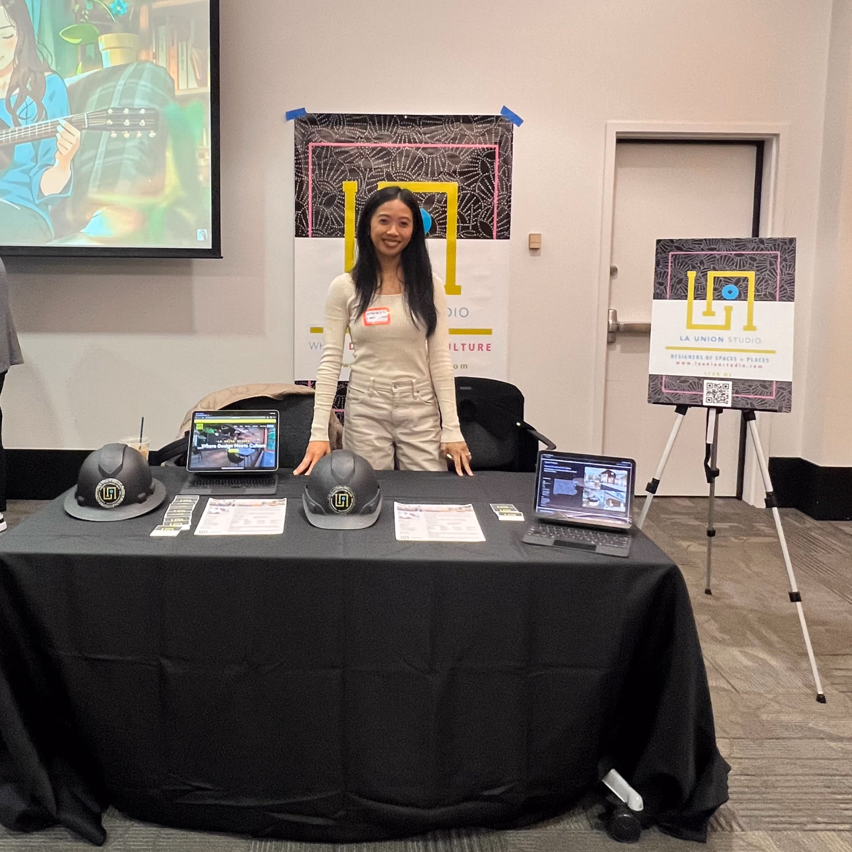 A person standing behind a black-draped exhibition table set with two laptops, printed handouts, and two branded hard hats, with a La Union Studio banner and easel poster behind them.