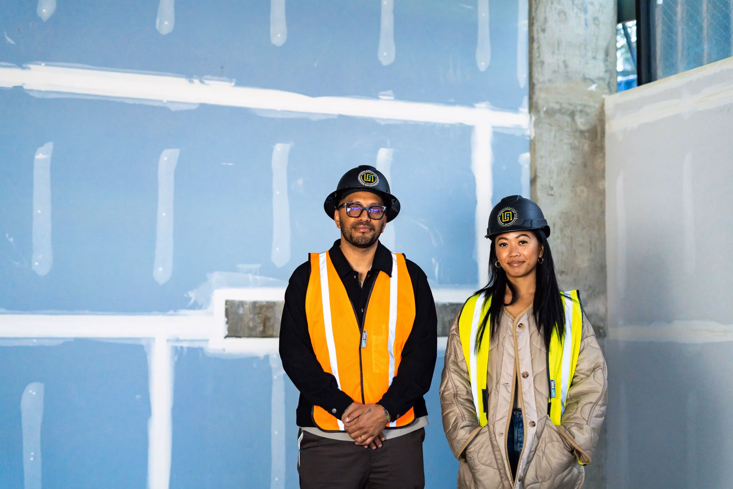 Sonia-Lynn and Sergio Max, founders of La Union Studio, standing in a construction site wearing safety vests and hard hats in front of a blue-painted framed wall