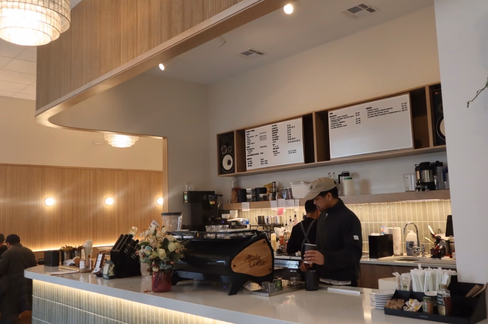 Two baristas working behind the curved counter of Ayala Coffee, with wood shelving, pendant lighting, and a menu board in the background