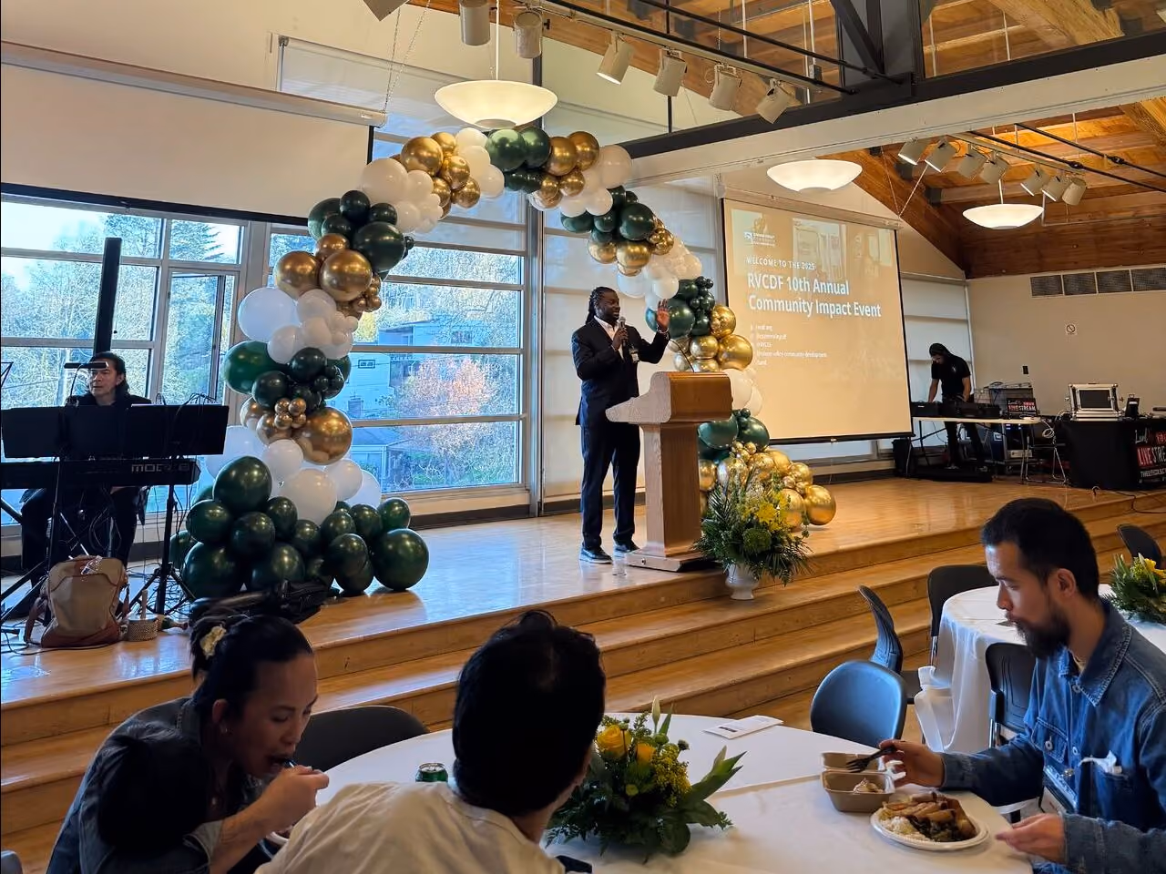 Speaker at a podium addressing an audience seated at round tables during the RVCDF 10th Annual Community Impact Event, with a gold and green balloon garland arching over the stage and a musician playing keyboard in the background