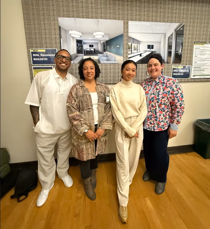 Four people posing together and smiling in a room with wood flooring, in front of a wall displaying interior renderings and community bulletin postings