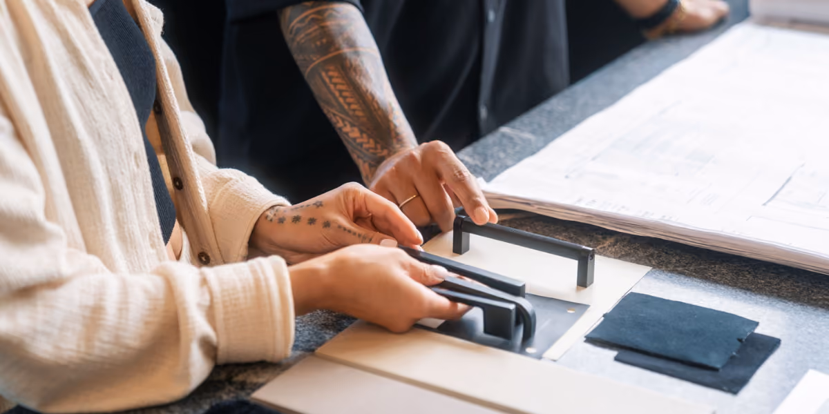 Close-up of two people's hands selecting cabinet hardware samples over architectural drawings on a table