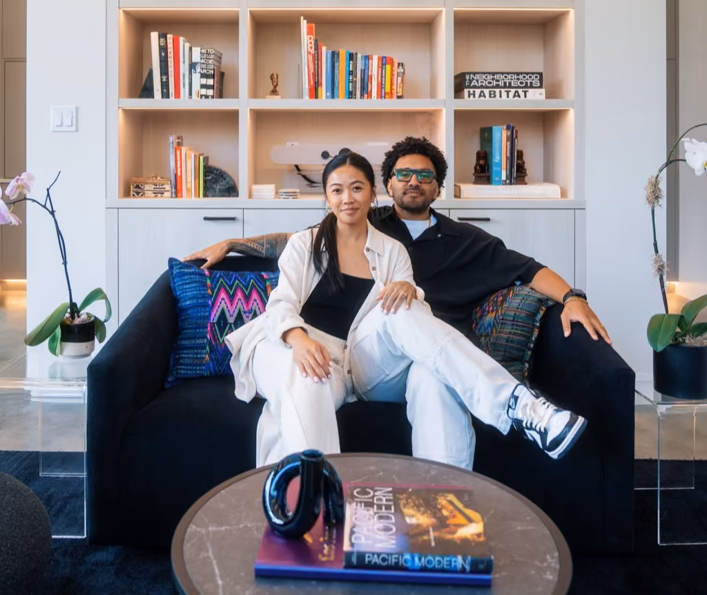 Sonia-Lynn and Sergio Max, founders of La Union Studio, sitting together on a black sofa in front of built-in shelving styled with books and decor, with a coffee table displaying the book 'Pacific Modern