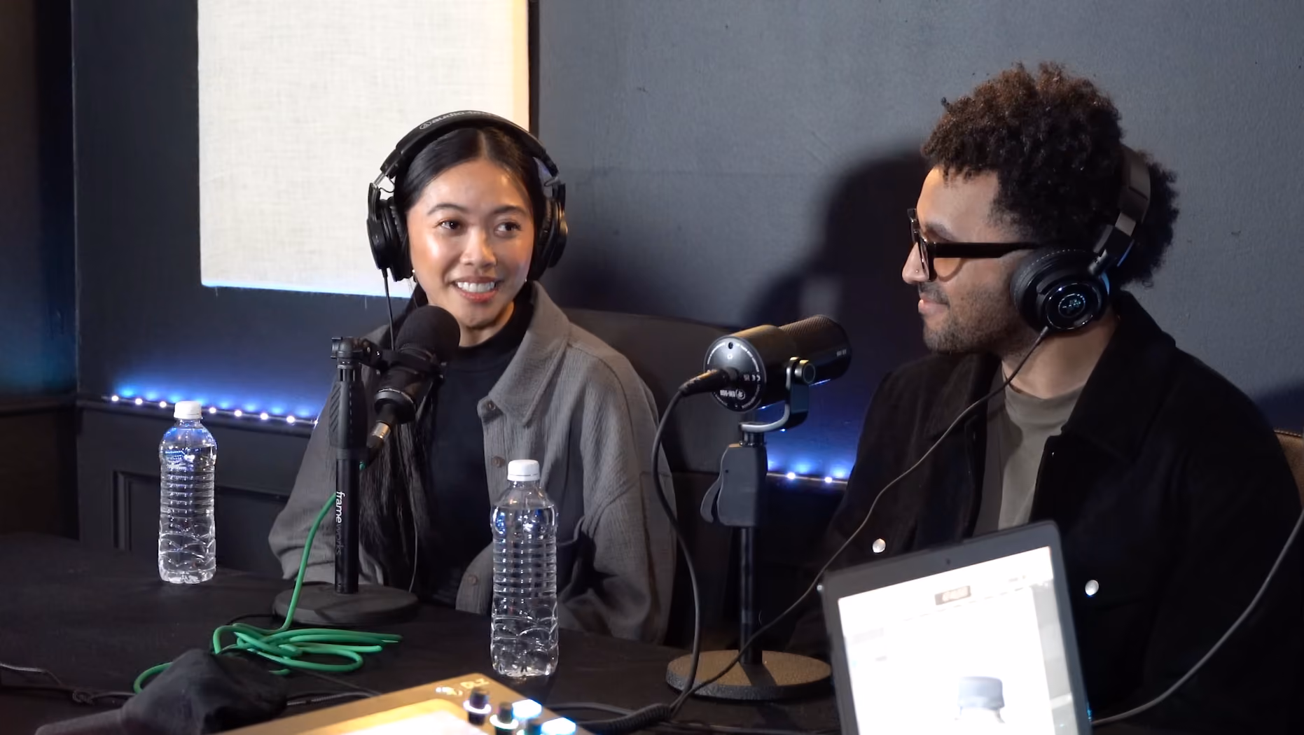 Woman and man sitting at a podcast recording setup wearing headphones, with microphones and water bottles on the table