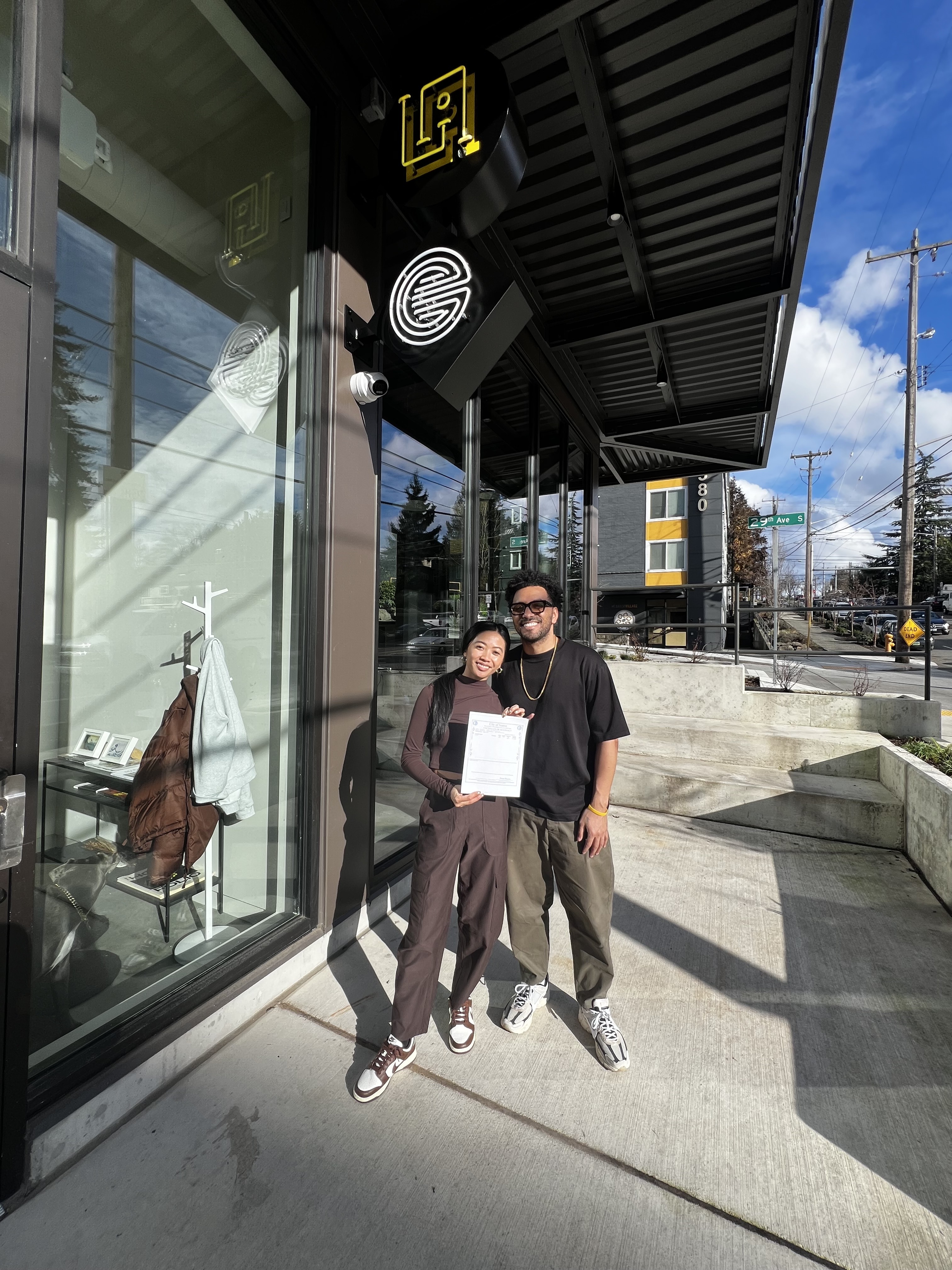 Two people standing outside a storefront holding a certificate, with skincare-related signage in the window