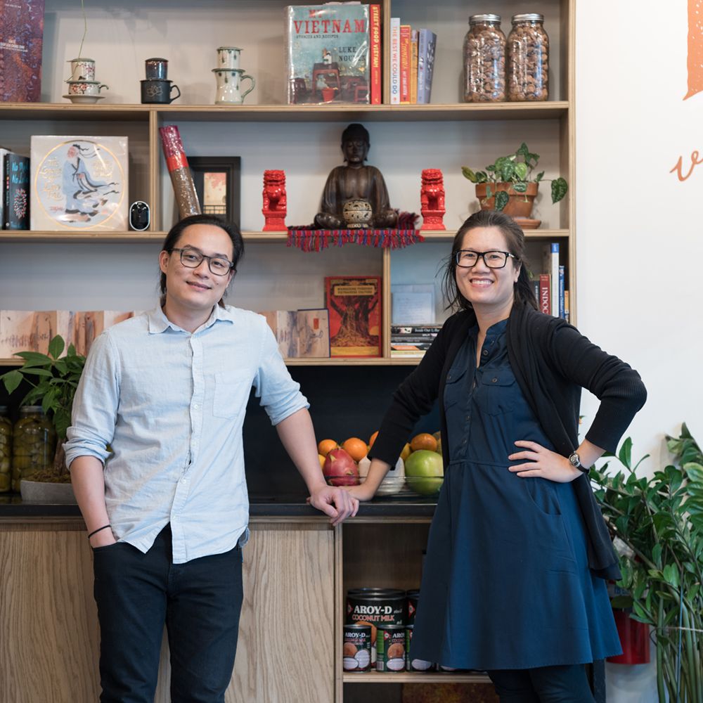 Two people standing side by side in front of the shelving at Phin Vietnamese Coffee, which holds books, a Buddha figure, bags of coffee, and a bowl of fruit on the counter below.