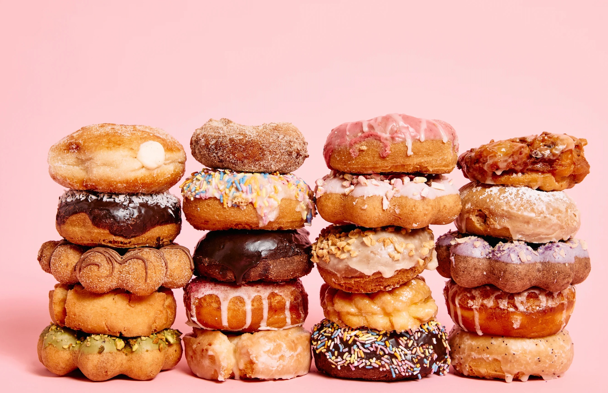 Four tall stacks of assorted doughnuts — glazed, chocolate-frosted, sprinkled, sugared, and filled — arranged against a plain pink background.
